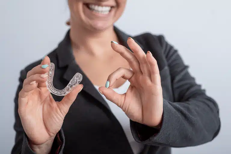Close-up of a smiling woman in business attire holding a clear dental aligner, showing orthodontic treatment option.