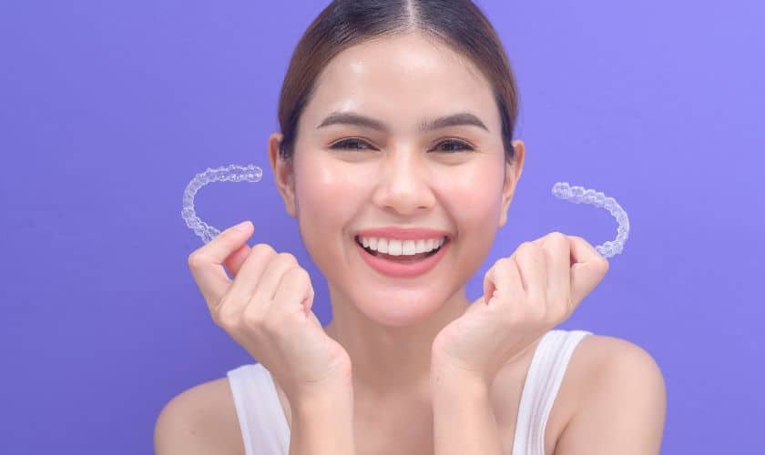 A smiling woman holding two Invisalign clear aligners in front of her face, showing excitement before starting orthodontic treatment.